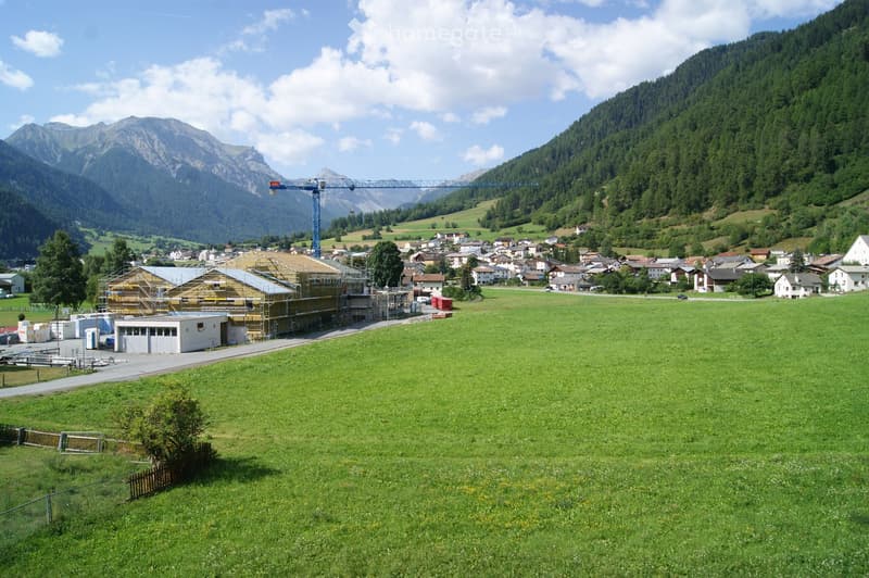 A rural village with a construction site, surrounded by green fields and mountains under a partly cloudy sky.
