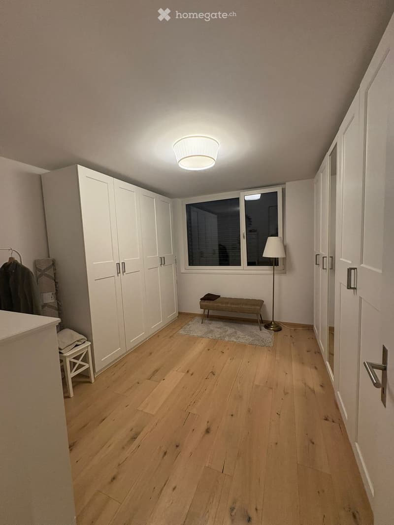A hallway with wooden flooring, white walls, and built-in wardrobes. A small bench and floor lamp are placed near a window, and a stool is visible on the left side.