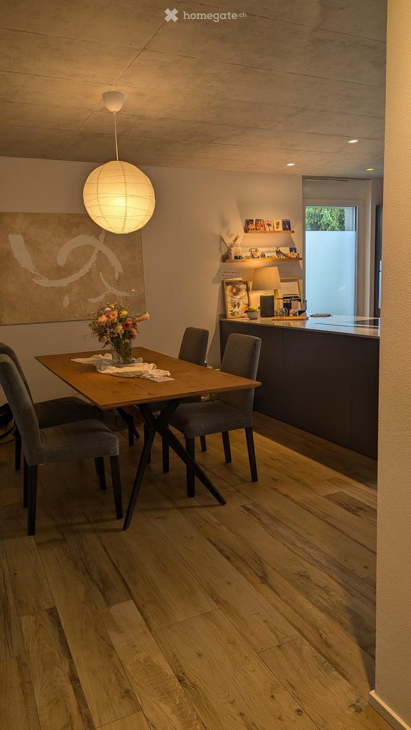 Modern kitchen and dining area with wooden flooring, a round pendant light, a wooden dining table with four chairs, and a vase of flowers on the table. The kitchen features a dark countertop with various items, including a lamp and framed pictures.