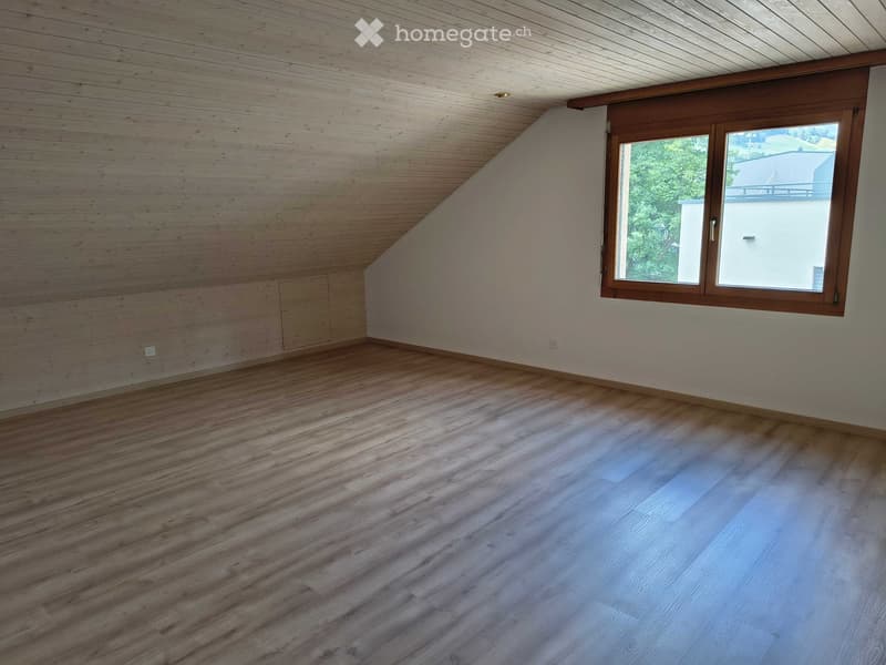 Empty room with wooden flooring, slanted wooden ceiling, and a large window with wooden frame. Bare walls, no furniture or equipment.