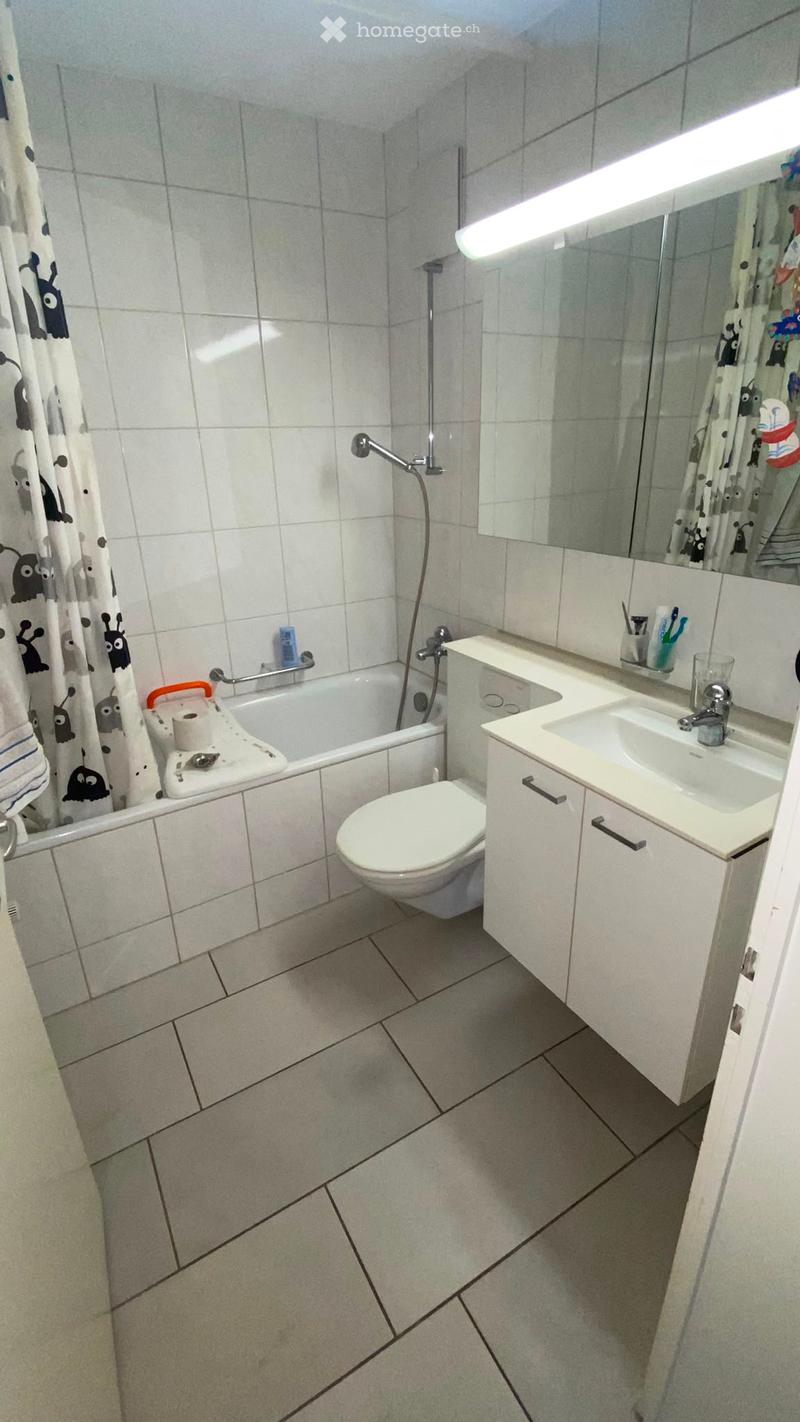 Modern bathroom with white tiles, a bathtub, toilet, sink, and mirror. Includes a shower curtain and a small cabinet with a mirror above the sink.