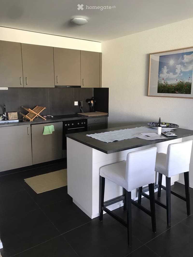 Modern kitchen with grey cabinets, black countertop, island with white stools, sink, oven, and a picture frame on the wall.