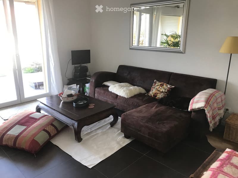 Living room with a dark brown sofa, coffee table, ottoman, mirror, television, and floor lamp. The room features large windows with white curtains and a view of the outdoors.
