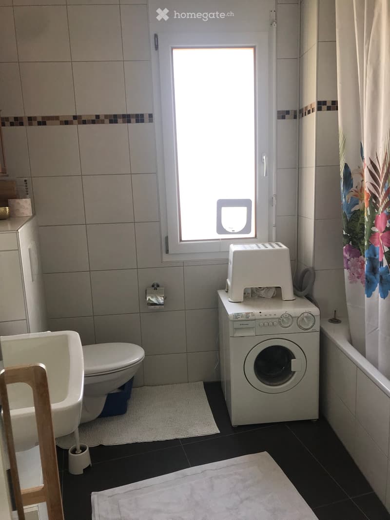 Bathroom with a toilet, washing machine, stool, and a small window. The room features tiled walls and a dark tiled floor with towels placed on the floor.