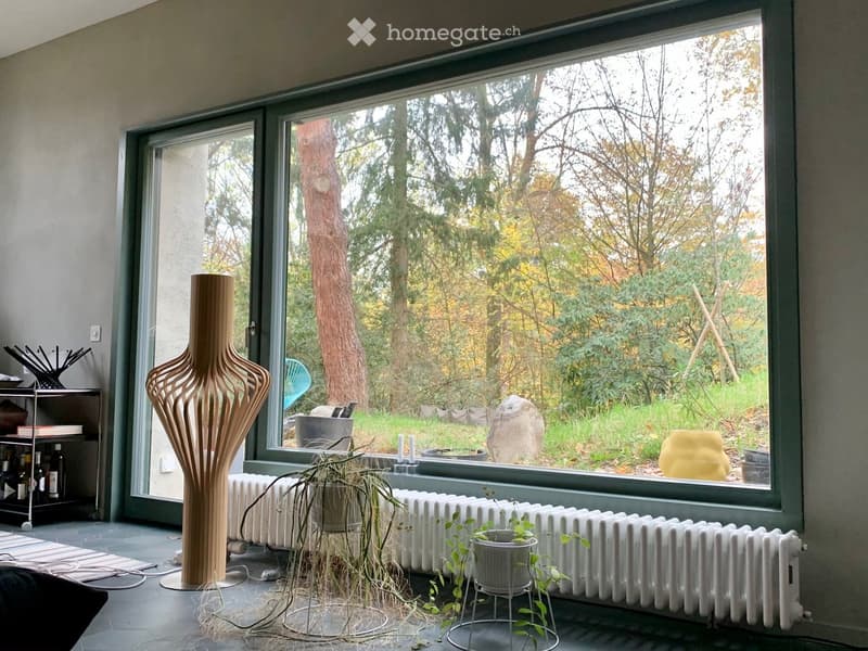 Modern living room with large windows overlooking a garden, featuring a tall wooden vase, radiator, potted plants, a shelf with bottles, and a small stool.