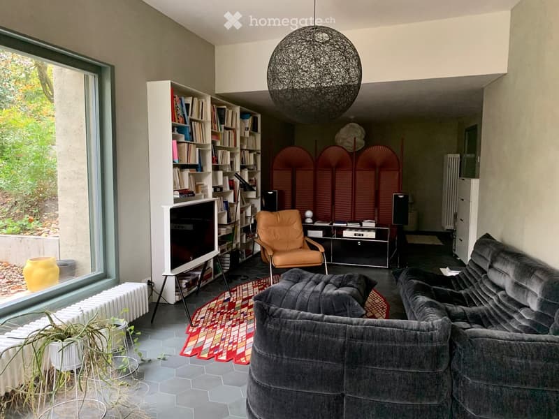 Modern living room with grey sectional sofa, orange armchair, bookshelf, TV screen, decorative lamp, and a view of greenery outside.