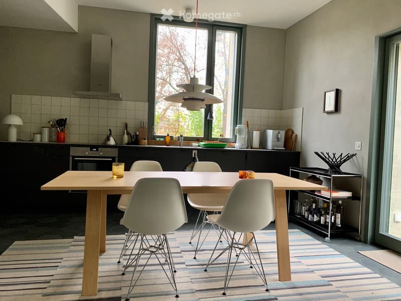 Modern kitchen with a large wooden dining table and four chairs, a built-in oven, a blender, and various kitchen items on the counter.