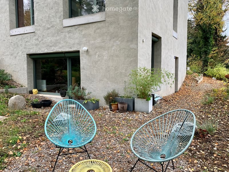 Modern outdoor seating area with two turquoise wire chairs and a yellow wire chair on a gravel surface, surrounded by potted plants and a concrete house facade.