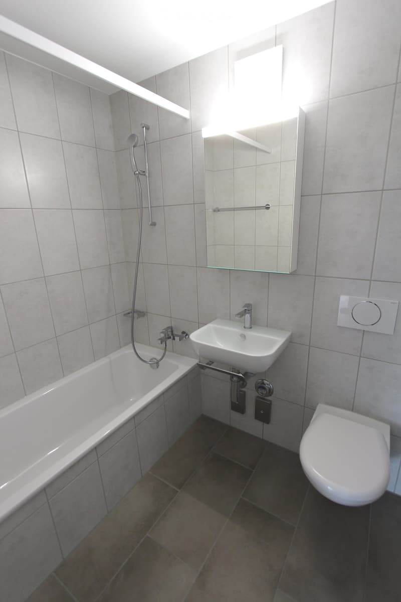 Modern bathroom with a bathtub, sink, toilet, and a large mirror with built-in lighting. The walls are tiled in light gray, and the floor is covered with dark brown tiles.