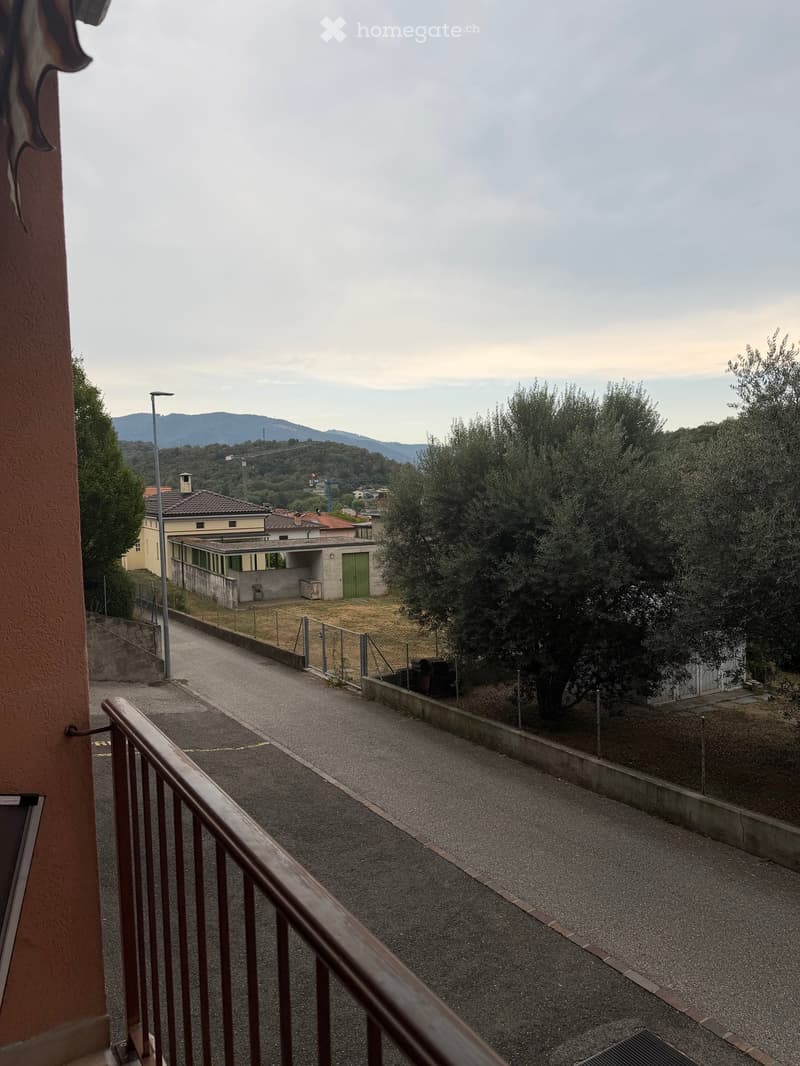 Balcony overlooking a paved pathway bordered by a low concrete wall, with trees and distant hills visible in the background.