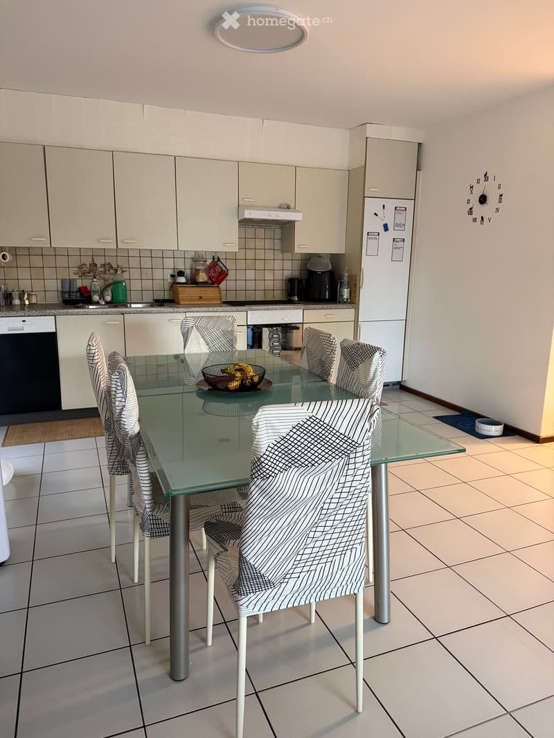 Modern kitchen with white cabinets, a glass dining table with six chairs, and various appliances including a refrigerator, dishwasher, and oven. The floor is tiled, and there is a bowl of fruit on the table.