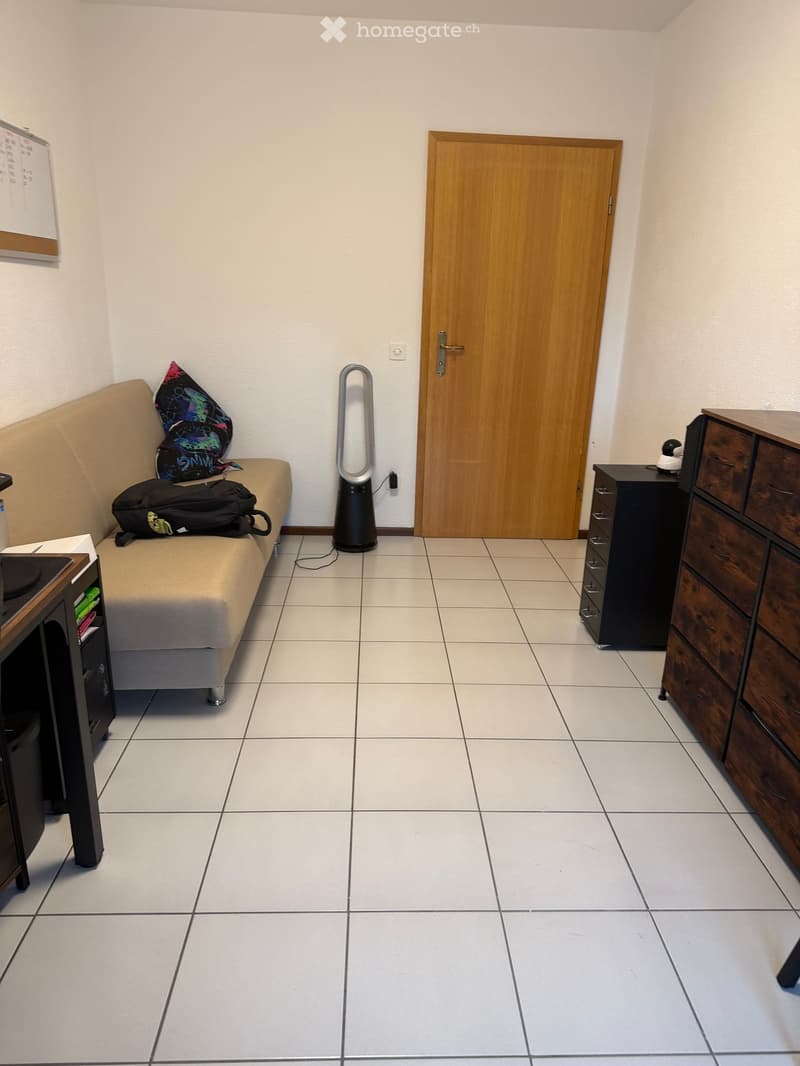 A small, furnished room with a sofa, a fan, a drawer unit, and a table. The room has tiled flooring and a wooden door.