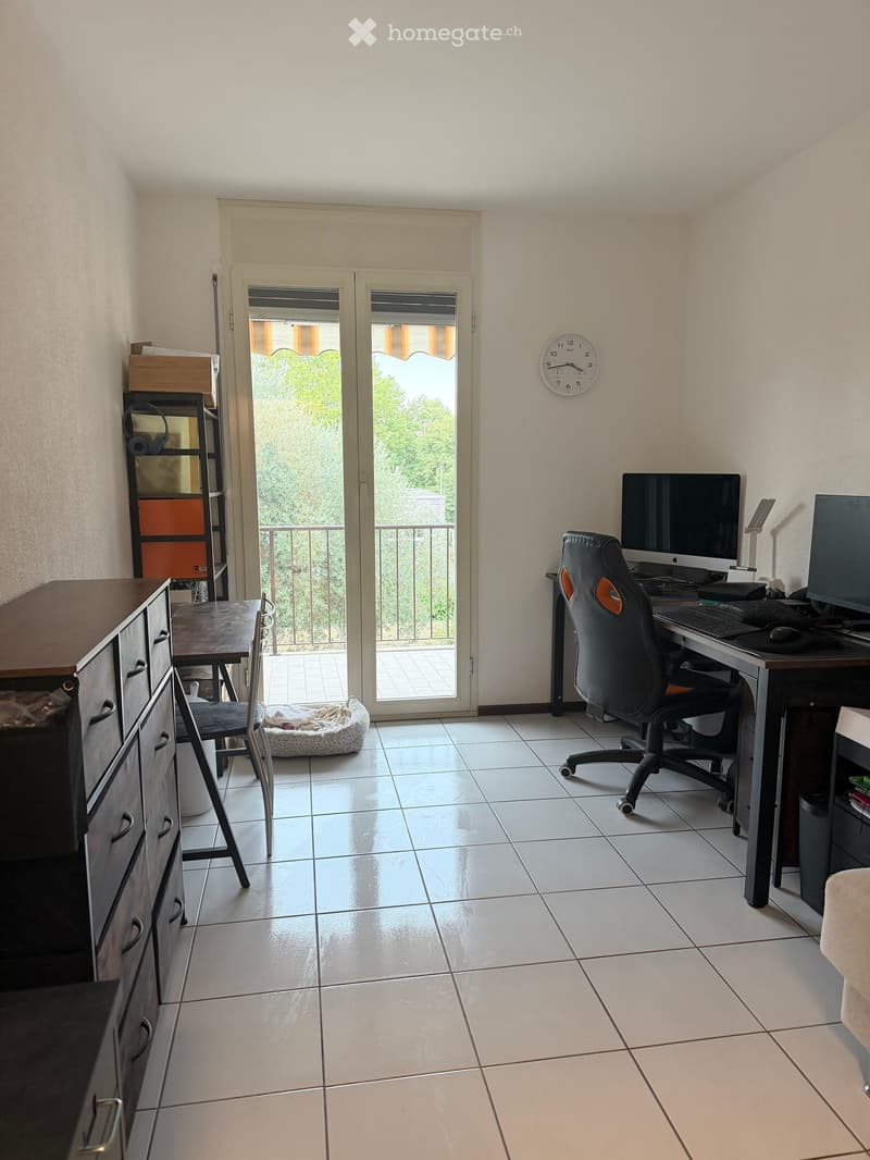 A furnished room with a desk, chair, screen, keyboard, mouse, clock, table, and shelf, featuring a balcony with a view of greenery.