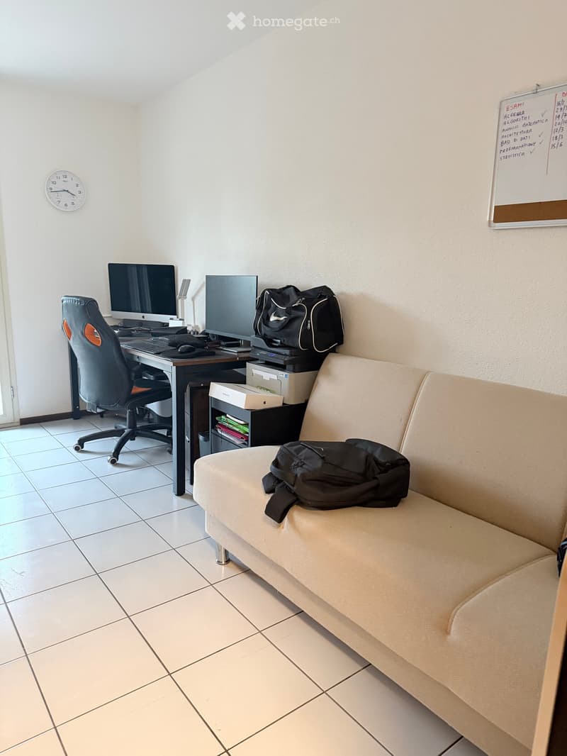 A furnished room with a sofa, desk, computer, chair, and a backpack on the sofa.