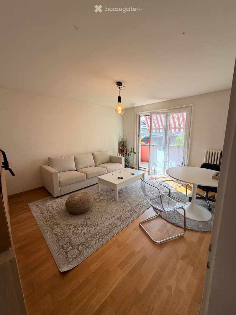 Living room with a beige sofa, white coffee table, round white dining table, modern chairs, and a patterned rug on wooden flooring.