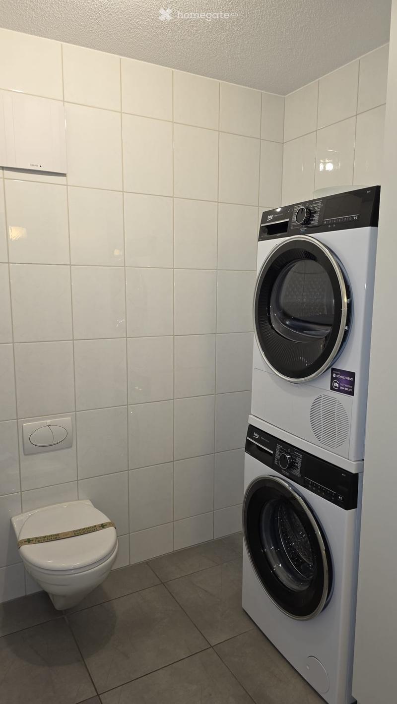 Bathroom with a toilet, washing machine, and dryer. The walls are tiled in white, and the floor is tiled in beige.