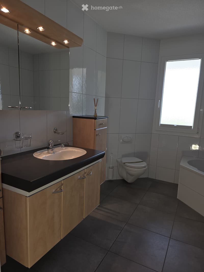 Modern bathroom with a sink, mirror, toilet, and bathtub. Features wooden cabinets, a black countertop, and a frosted window.