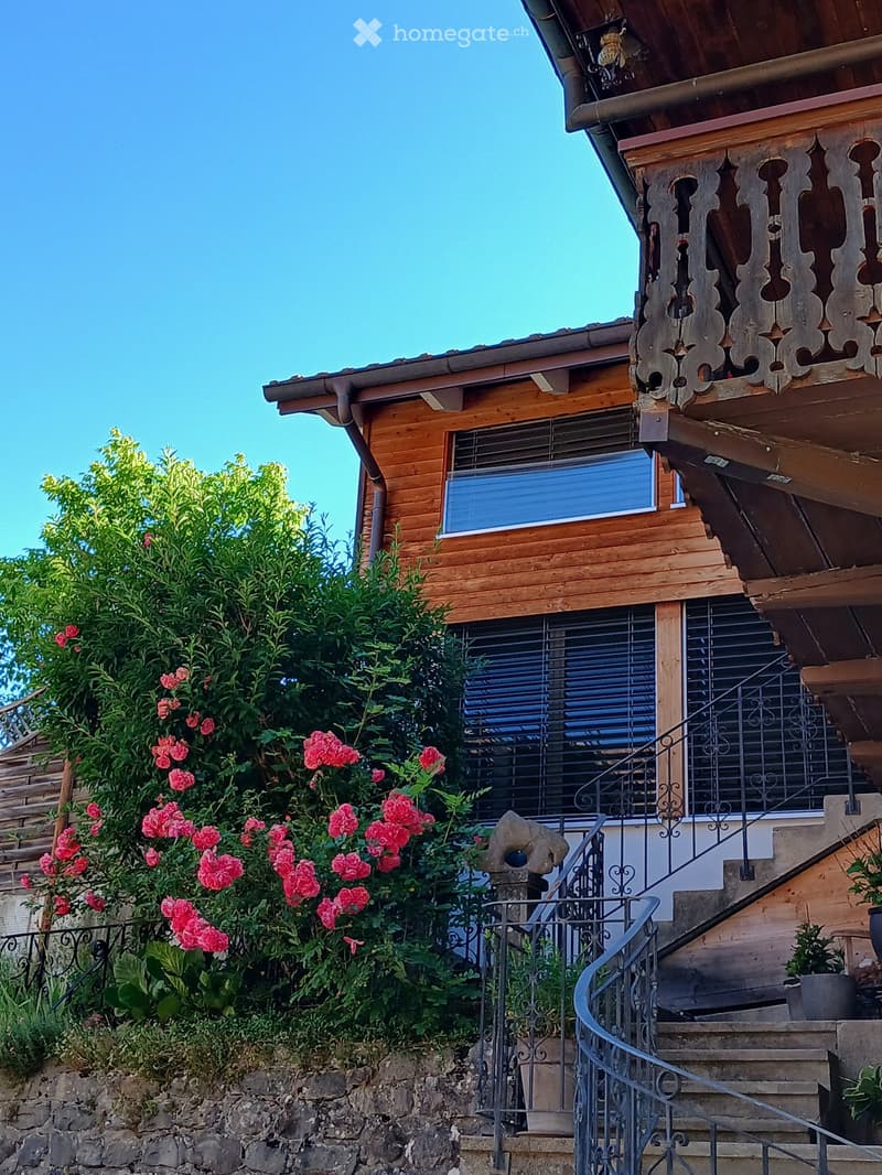 Two-story wooden house with a balcony, ornate wooden railing, and a staircase leading up to the entrance. The house has multiple windows with blinds and is surrounded by greenery and pink flowers.