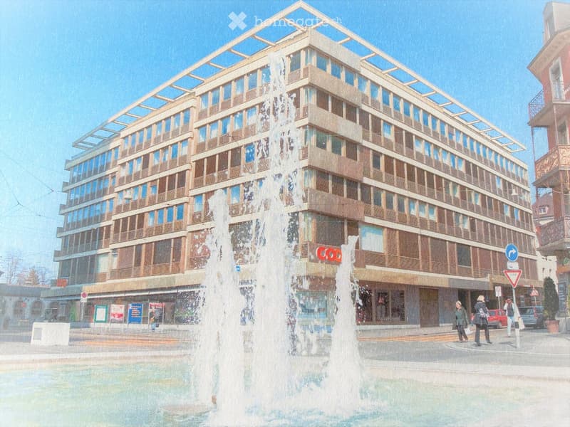 Modern multi-story building with large windows and balconies, located in an urban area with a fountain in the foreground.