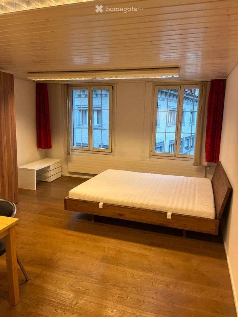 A modern, minimalist bedroom with wooden flooring, a bed with a white mattress, a small storage unit, a chair, and a table near the window.