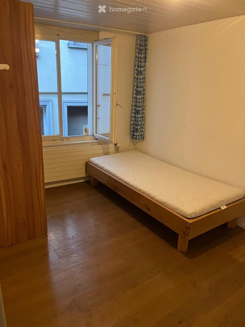 Small bedroom with wooden floor, single bed, open window with view, and a curtain.