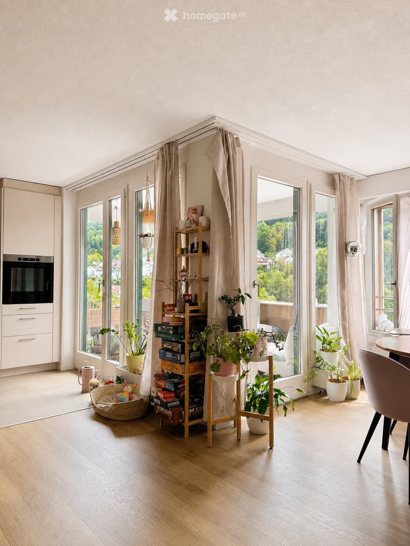 Bright, airy room with large windows and a view of greenery, featuring a wooden floor, a shelf with books and plants, a wicker basket, and a small table with a potted plant.