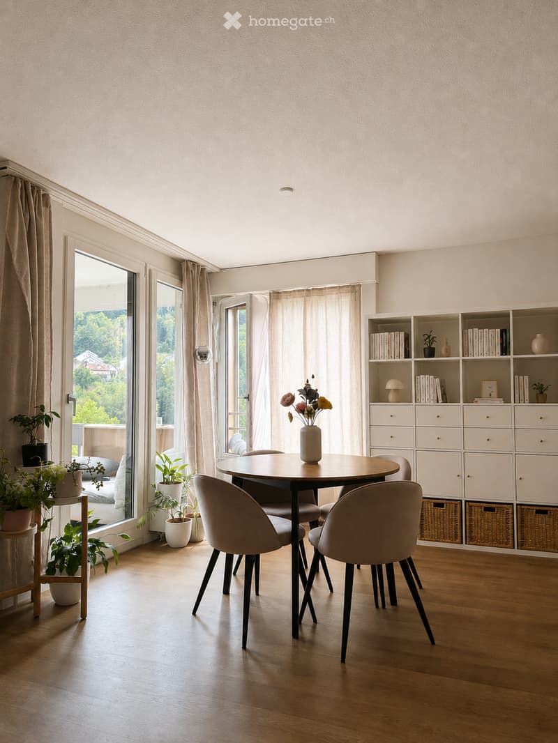 Modern dining area with a round wooden table, four chairs, a bookshelf, and potted plants near large windows with curtains.