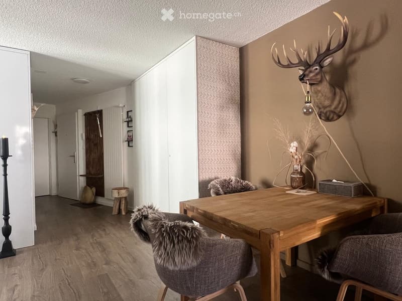 A modern living room with a wooden table, two chairs, a candle, a vase with dried flowers, a deer head on the wall, and a small stool near the door.