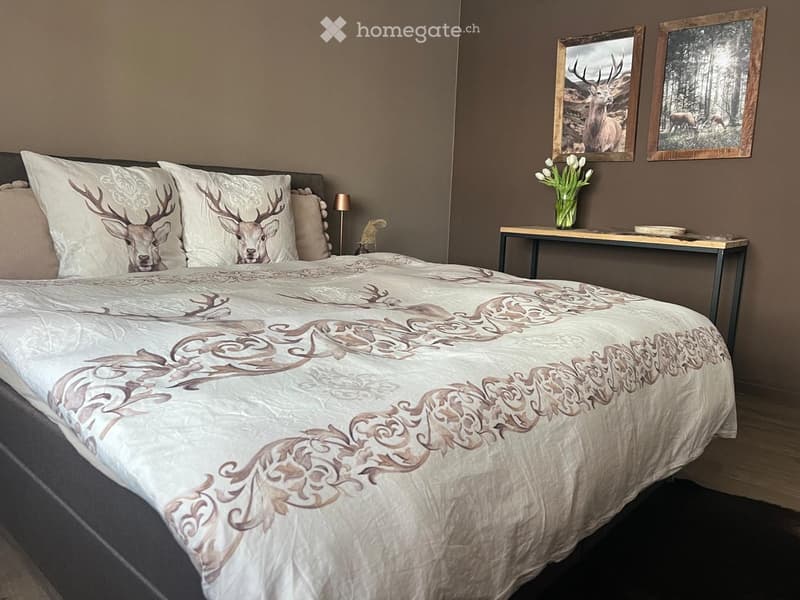 A well-furnished bedroom with a large bed, decorative pillows, a nightstand, a small table with a vase of flowers, and framed artwork on the wall.
