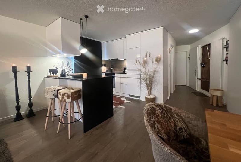 Modern kitchen with white cabinets, black island, and wooden flooring. Features two stools at the island, a fur throw on a chair, and decorative candles.