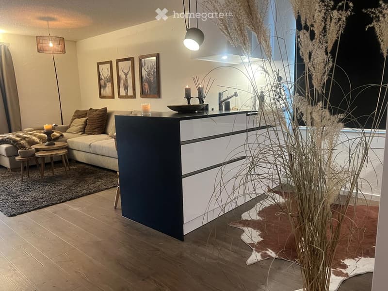 Modern living room with a sectional sofa, coffee table, floor lamp, and decorative items. Features a sleek black and white bar area with a faucet and candle, along with framed artwork and a tall plant in the corner.
