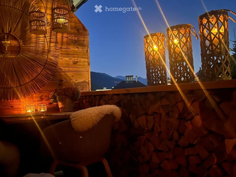 Cozy balcony with wooden walls, decorative lanterns, a small table with candles, a chair with a fur throw, and a stack of firewood.