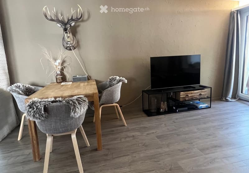 Modern living room with wooden flooring, a large flat-screen TV on a black stand, a wooden dining table with four chairs, a decorative deer head on the wall, and a small table lamp.