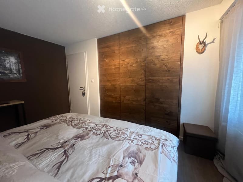 Bedroom with a large bed, wooden wardrobe, small stool, and a mounted deer head on the wall.