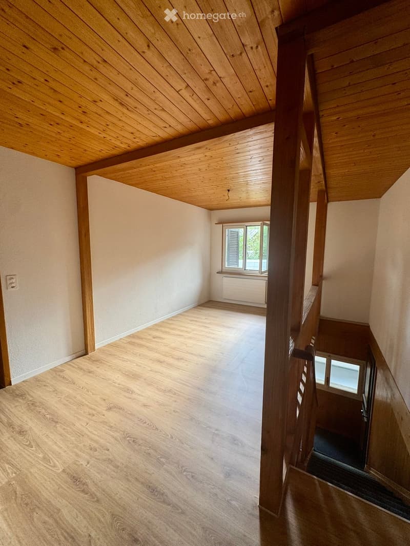 Empty room with wooden flooring and light-colored walls, featuring a staircase leading upwards and a window with shutters.