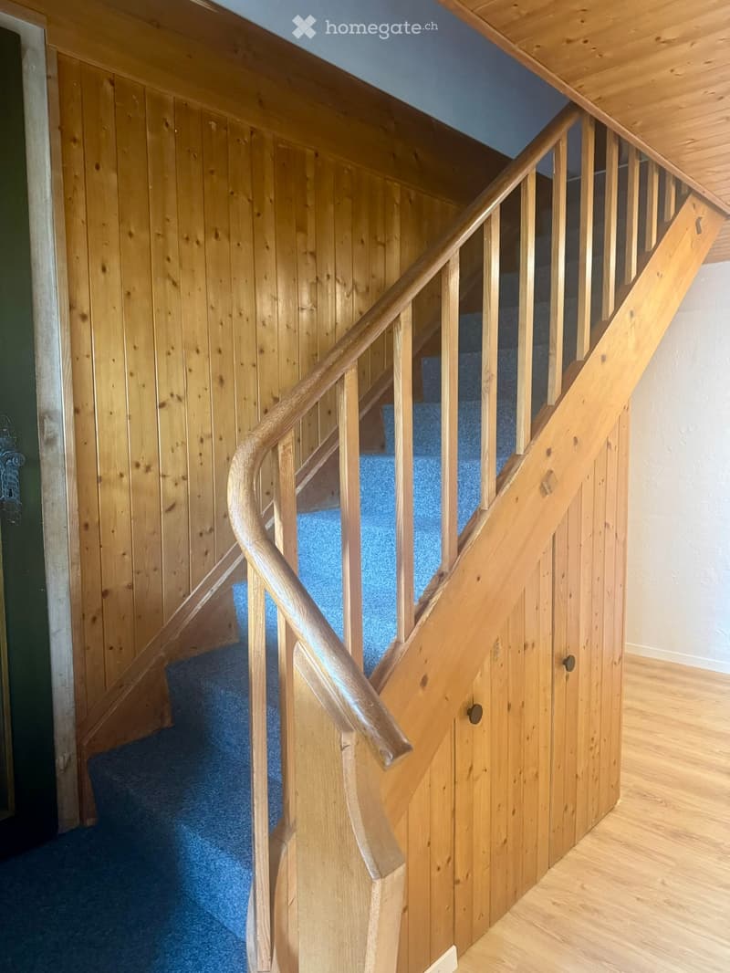 Wooden staircase with a curved handrail and blue carpeted steps, leading to an upper level.