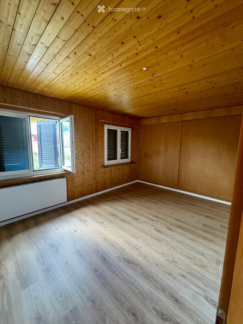 Empty room with wooden walls and ceiling, wooden flooring, and two windows, one of which is open.