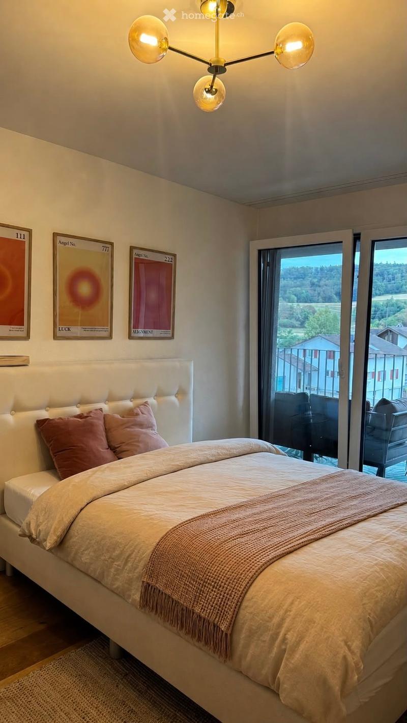 Modern bedroom with a large bed, decorative pillows, a cozy blanket, wall art, and a view through large windows.