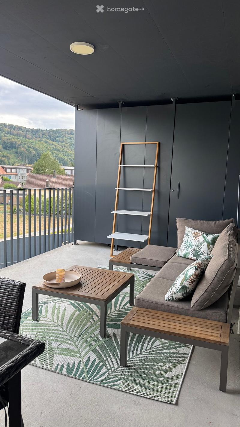 Modern balcony with grey sectional sofa, wooden coffee table, wicker chair, and a shelf unit, overlooking a green landscape.