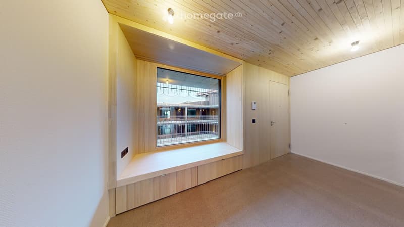 Empty room with wooden ceiling, white walls, carpeted floor, built-in bench seating, and a large window with a view of an outdoor area.