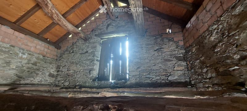 Empty room with exposed wooden beams and stone walls, featuring a wooden door and small windows allowing natural light to enter.