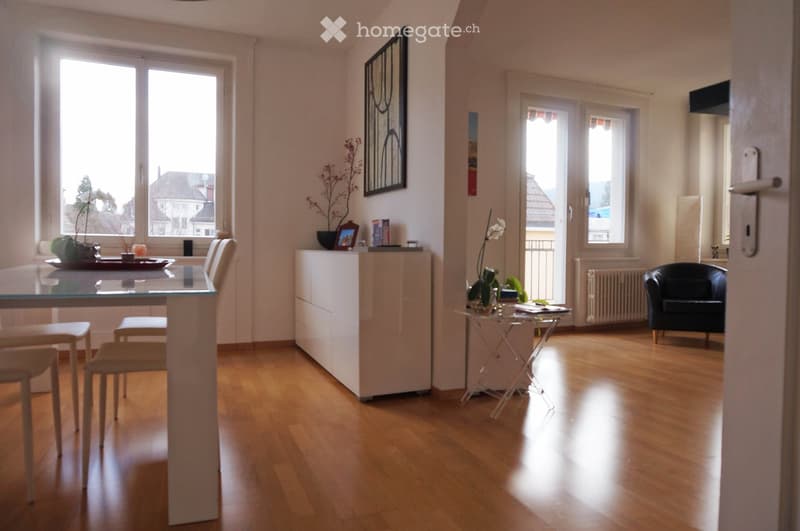 Bright, open living space with wooden flooring, large windows, a glass dining table with chairs, a white cabinet, a black armchair, and a small side table with a vase of flowers.