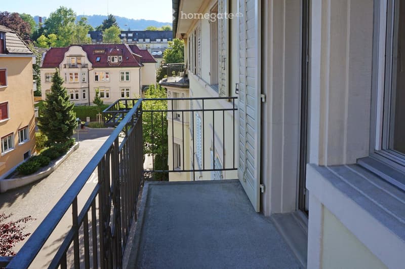 Balcony with metal railing, overlooking a street lined with residential buildings and greenery.