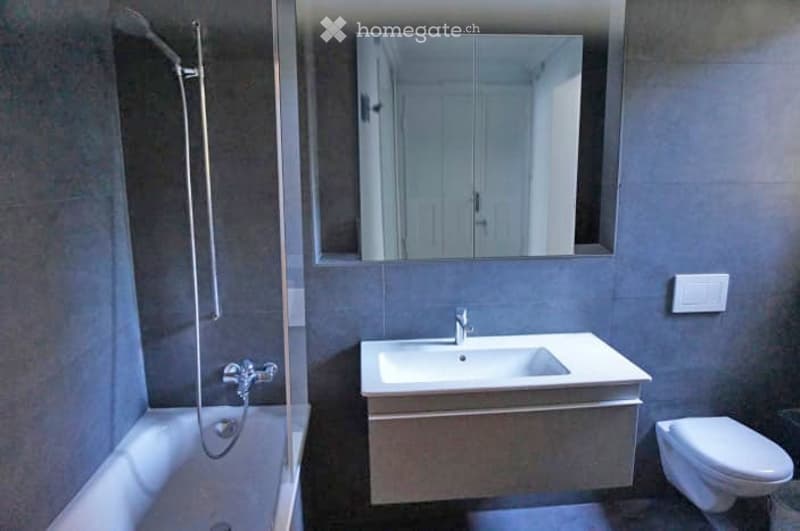 Modern bathroom with a white sink, mirror, toilet, and bathtub. Grey tiles and sleek fixtures.