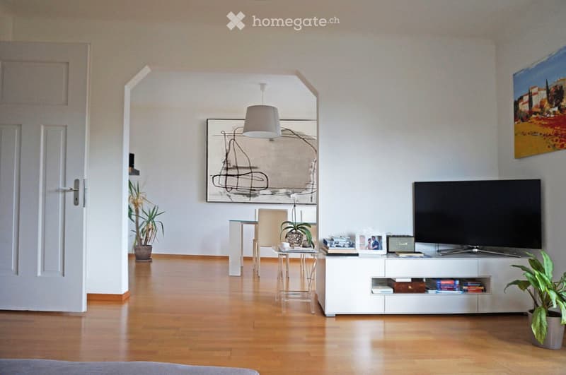 Modern living room with wooden flooring, white walls, and large windows. Features a television on a white stand, a small table with a chair, potted plants, and artwork on the walls.