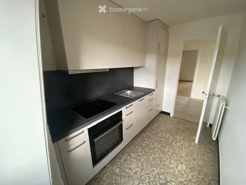 Modern kitchen with white cabinets, dark countertop, built-in stove and oven, and a sink. The floor is tiled, and there is an open doorway leading to another room.