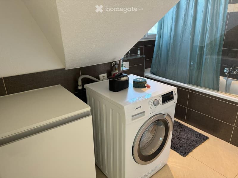 Bathroom with a washing machine, a bathtub, a mirror, and a small cabinet. The walls are tiled in dark brown and white, and there is a window with a blue curtain.