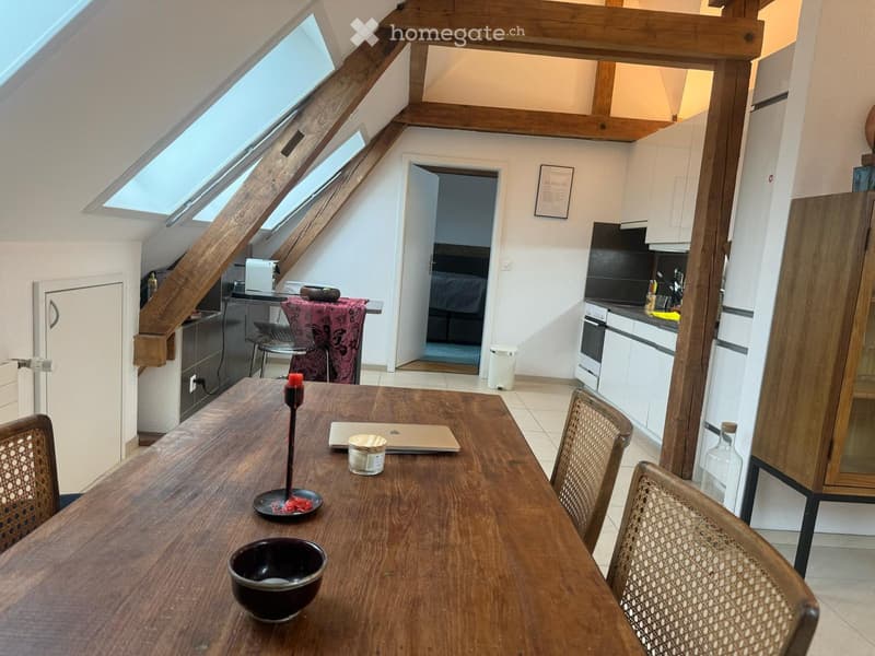 A modern, well-lit room with wooden beams and skylights, featuring a dining table with chairs, a laptop, and a kitchen area with white cabinets, a refrigerator, and an oven.