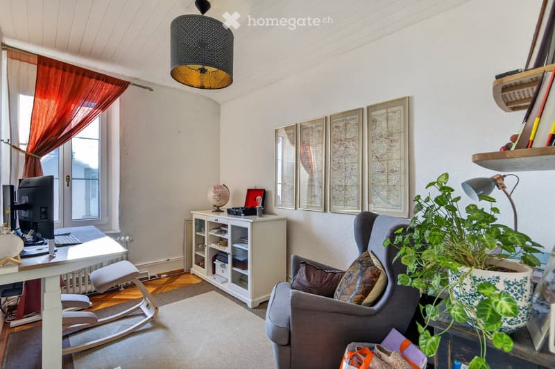 A cozy, well-furnished room with a grey sofa, armchair, white desk, computer, hanging lamp, plant, and decorative items including framed maps and a globe.