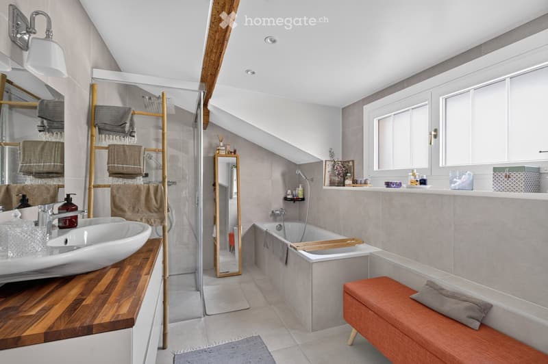 Modern bathroom with a white sink, bathtub, wooden stool, mirror, towel rack, and shelves. The space features large windows and a sleek design with neutral tones.